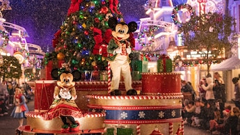 Mickey Mouse y Minnie Mouse vestidos con atuendos festivos en una carroza decorada con un árbol de Navidad gigante y regalos