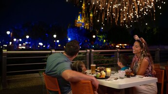 A family of 4 dine on an outdoor terrace as fireworks burst above Cinderella Castle in the background