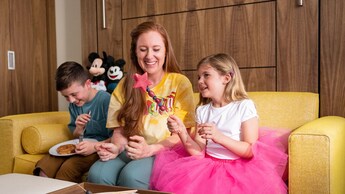 Una madre y dos niños sentados en un sofá junto a dos peluches de Mickey Mouse, comiendo frutas y panqueques