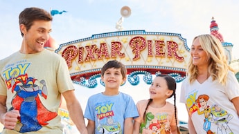 A family of 4, wearing T shirts illustrated with Disney characters and text that reads 'Toy Story,' stands near a sign that reads 'Pixar Pier'