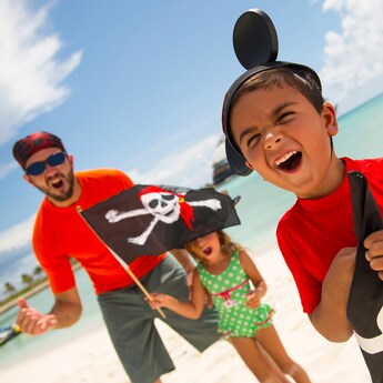 A father wearing a bandana, a girl holding a Jolly Roger flag and a boy with a Mickey Mouse ear hat show off their best scowls on the shores of Disney Castaway Cay
