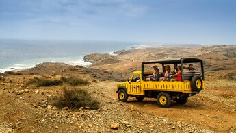 Una Land Rover Defender a cielo abierto, llena de pasajeros, se detiene en un terreno rocoso no desarrollado con vista al océano