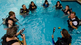 Un grupo de estudiantes de buceo está de pie en una piscina y se reúne alrededor de un instructor de buceo mientras aprende a utilizar sus equipos