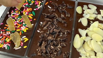 A hand adding various toppings to chocolate bars in a molding tray