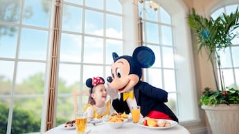 Mickey Mouse whispering something into a smiling young girl’s ear at a breakfast table