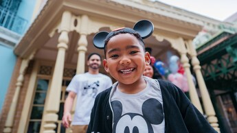 A young boy smiling and standing in front of his parents while wearing a Mickey Mouse ear hat and shirt