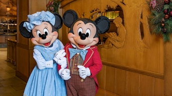 Minnie Mouse and Mickey Mouse standing arm in arm while wearing holiday attire