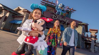 A young girl wears a Minnie Mouse ear headband and hugs a Minnie Mouse plush while walking with her parents near the World of Disney store