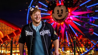 Guest wearing a Disneyland 70th anniversary baseball jersey and Mickey Mouse ear headband stands near a railing with a brightly lit Ferris wheel featuring a Mickey face behind them at night