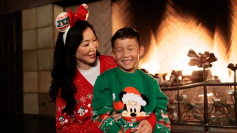 A mother wearing a holiday themed sweater and mouse ears holds her young son, who sports a sweater featuring Mickey Mouse in a Santa hat