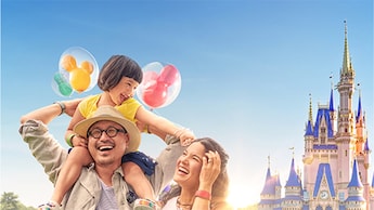 A family of 3 Guests including a daughter riding on her father’s shoulders while holding a bunch of Mickey Mouse balloons in front of Cinderella Castle