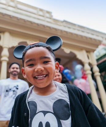 Un niño sonriente de pie frente a sus padres con un sombrero con orejas y una camisa de Mickey Mouse
