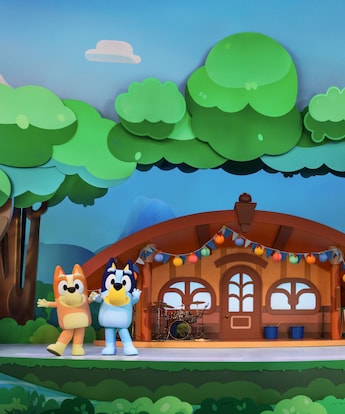 Bingo and Bluey pose on a stage set designed to look like a house in the woods at Bluey’s Best Day Ever