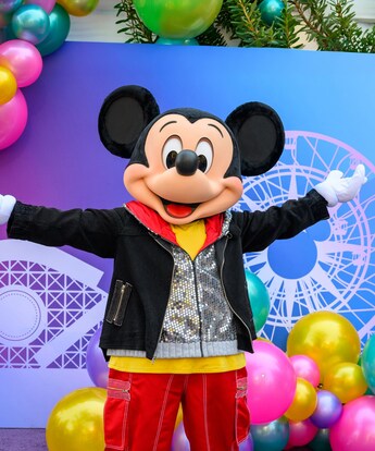 Mickey Mouse standing with arms outstretched near a photo op wall with balloons