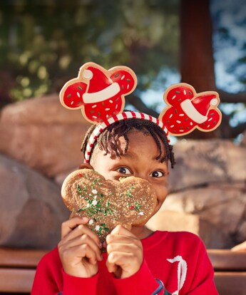 A young girl in Christmas themed Minnie ears holds up a big Mickey Mouse shaped holiday cookie