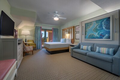 A Disney’s Vero Beach Resort hotel room with a sofa, bed, 2 wall sconces, side table, chairs, lamp, dresser, TV and aquatic artwork	