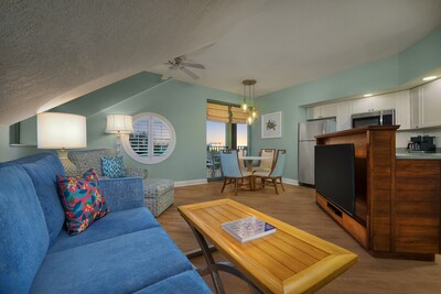 A room with a vaulted ceiling that includes a sofa, sleeper chair, side table, refrigerator, microwave, kitchen bar, TV and window with deck views	