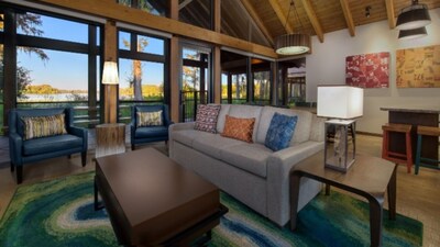 A common area in a 2 Bedroom Cabin at Copper Creek Villas & Cabins at Disney's Wilderness Lodge with a dining counter, 2 stools, a lamp table, a sofa, a coffee table, 2 armchairs and 4 windows with lakefront views
