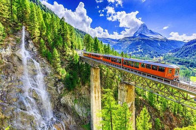 A train on the Gornergrat Railway near Zermatt, Switzerland passes a waterfall, with the Matterhorn in the distance