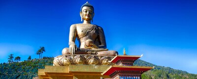 A massive Buddha statue with a bowl sits atop a building overlooking a small temple in Thimphu