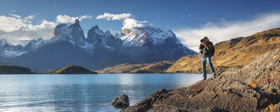 A man standing on a rocky hill overlooking a lake with nearby mountains takes a photo with a camera