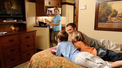 A father and 2 children lie on a bed watching TV while the mom pours popcorn into a bowl