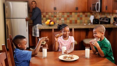Three children eat cookies at a dining room table while their father watches from the kitchen