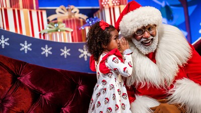 A child wearing a dress with the faces of Mickey Mouse and Minnie Mouse while whispering into Santa’s ear