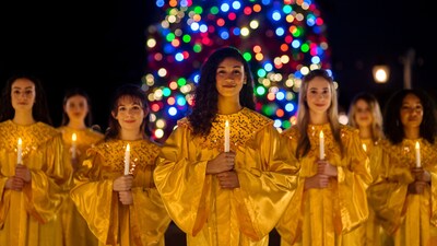 A choir group holds candles in front of an illuminated Christmas tree at night