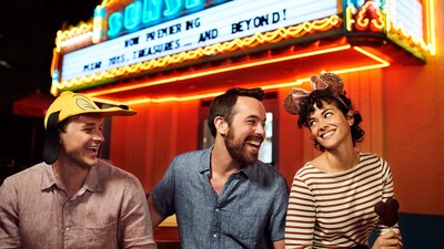 A group of friends where Disney hats while sitting outside of shop with a sign that says, ‘Sunset Now Premiering’