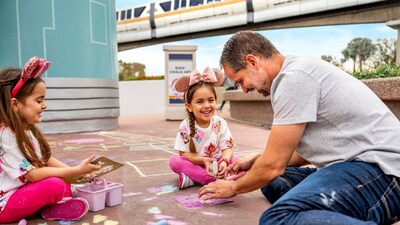 A man and 2 girls using colorful chalk to decorate a sidewalk near the monorail line