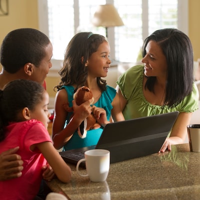 A man, woman and 2 daughters gathered around a laptop in their living room