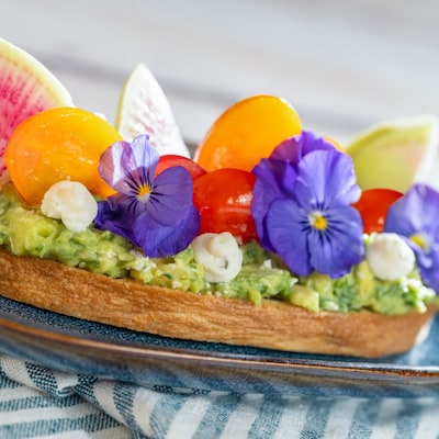 Uma fatia grossa de torrada com abacate, coberta com tomates-uva e enfeitada com flores frescas
