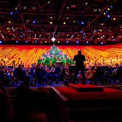 Una persona dirigiendo una orquesta y un coro; algunos miembros del coro están vestidos y dispuestos para verse como un árbol de Navidad en Candlelight Processional