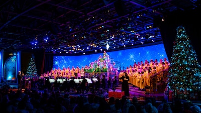A conductor leading a large choir and orchestra on a festive holiday stage flanked by decorated Christmas trees