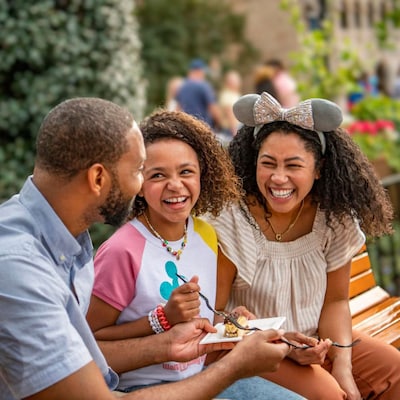 A man, woman and their daughter sitting on a park bench, sharing a snack