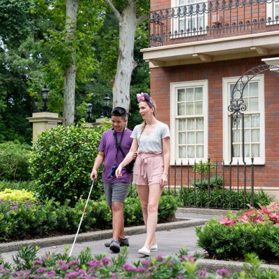 A boy with a white cane wears headphones and the Disney Handheld Device, while holding on to a woman’s arm.