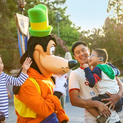 A father holds his son with a cochlear implant in his arms while laughing with Goofy