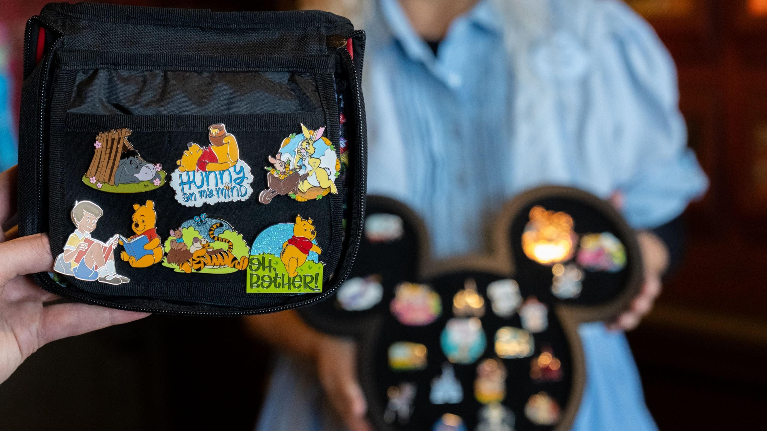 A hand holding a pin trading book with a Cast Member holding a Mickey Mouse shaped pin tray nearby
