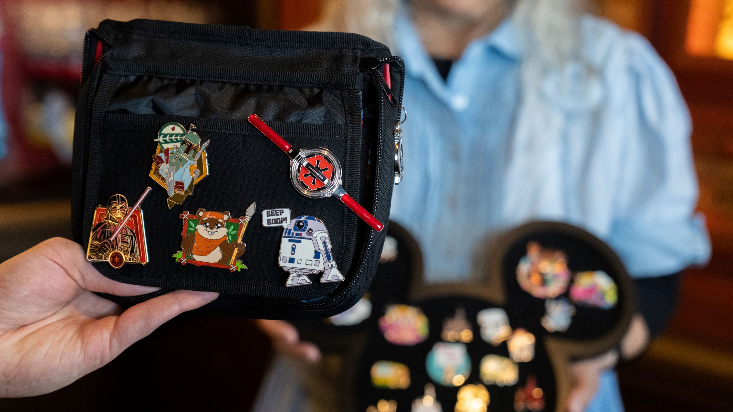 A hand holding a pin trading book with a Cast Member holding a Mickey Mouse shaped pin tray nearby
