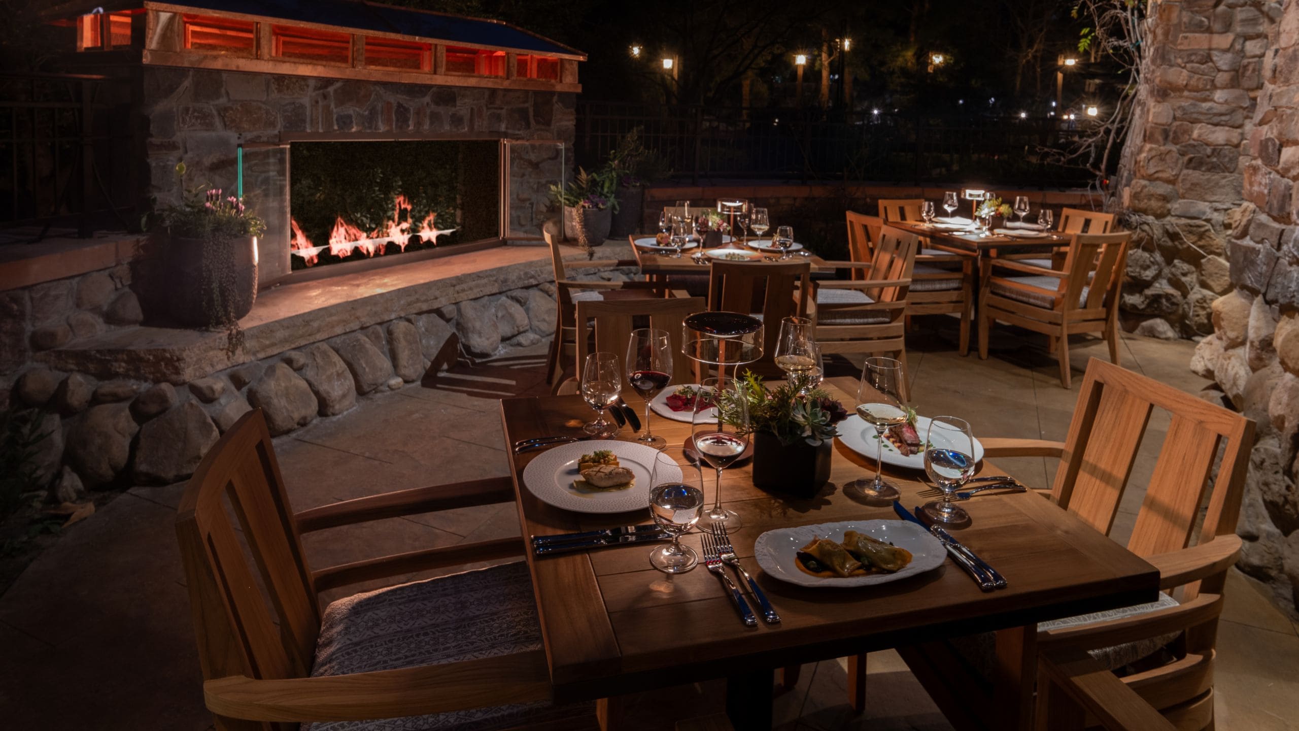 A terrace dining area in Napa Rose at night, with a stone fireplace and wooden tables and chairs