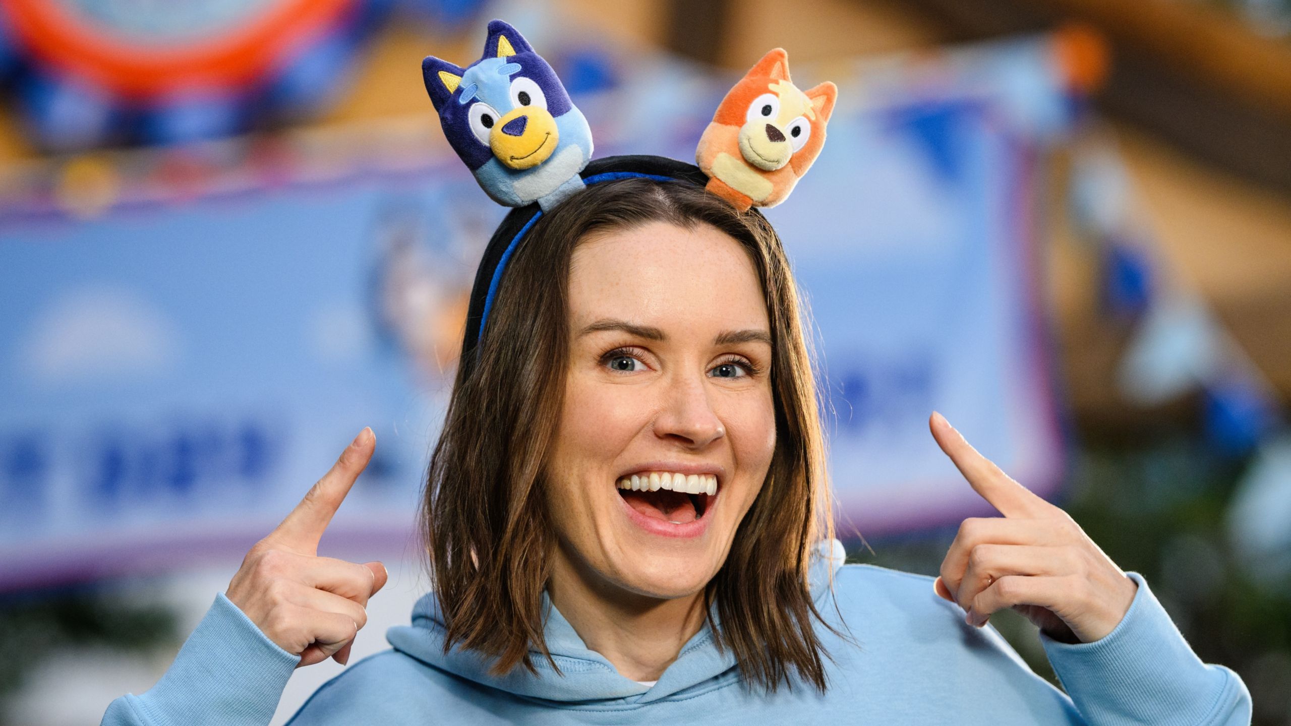 A woman pointing to a Bluey and Bingo plush headband, available during Bluey's Best Day Ever
