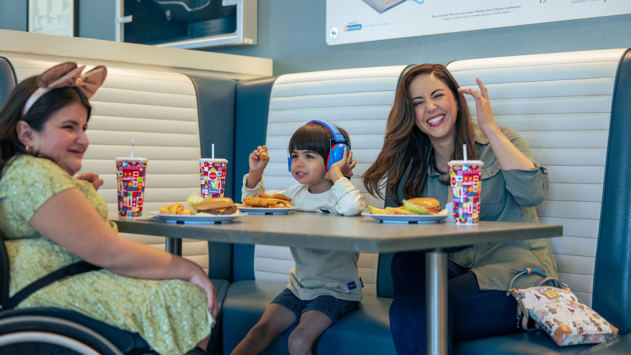 Una mujer en silla de ruedas, un niño con audífonos y una tercera mujer sentados en un restaurante disfrutando de hamburguesas y refrescos.