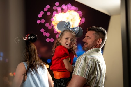 A father holds his daughter in his arms while they smile as fireworks burst behind them