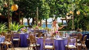 Outdoor event setup with round tables covered in linens and floral centerpieces with a Cast Member arranging decor