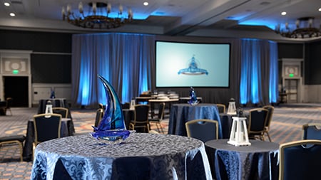A ballroom with set tables, chairs, decorative centerpieces and a projection screen flanked by curtains A ballroom with set tables, chairs, decorative centerpieces and a projection screen flanked by curtains