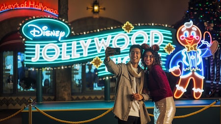 A couple takes a selfie in front of a neon sign that reads ‘Disney Jollywood Nights’ 