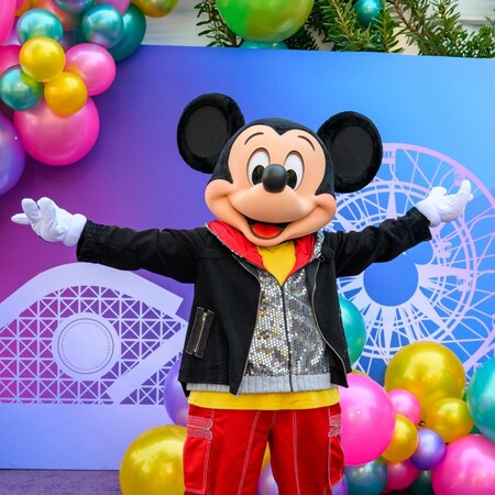 Mickey Mouse standing with arms outstretched near a photo op wall with balloons
