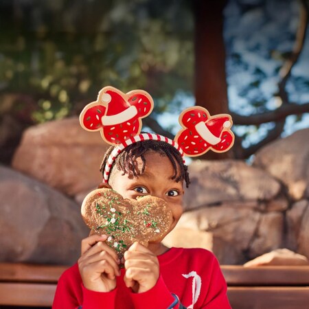 A young girl in Christmas themed Minnie ears holds up a big Mickey Mouse shaped holiday cookie