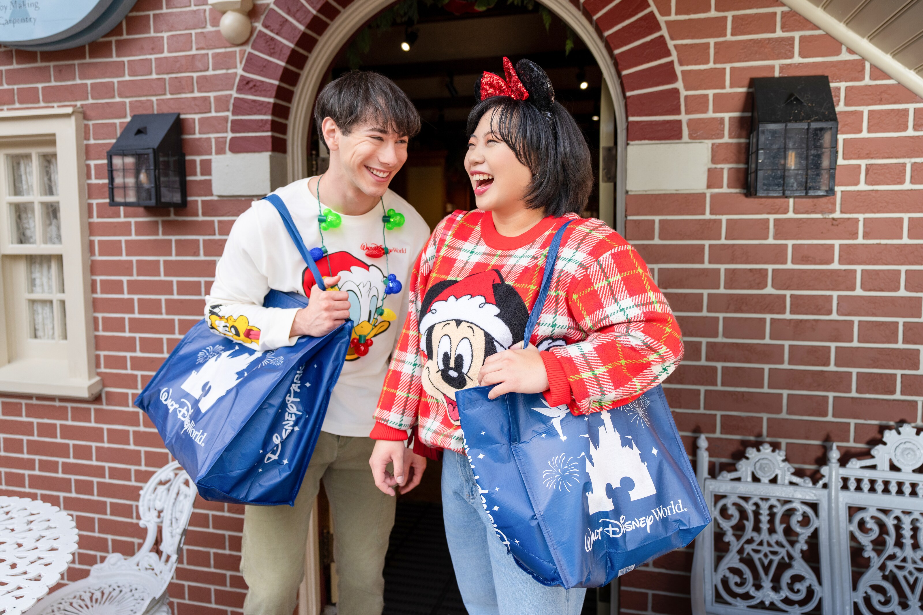 Two Guests wearing Christmas apparel including a sweater featuring Mickey Mouse, a long sleeve shirt featuring Donald Duck, Pluto and Goofy, a Minnie Mouse ear headband and a string light necklace